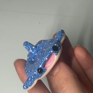 May include: A small, blue whale shark figurine featuring a pattern of white dots across its back, a smooth cream-colored underside, two black circular eyes, and a wide, open mouth with a pink interior. The shark has a dorsal fin on its back.