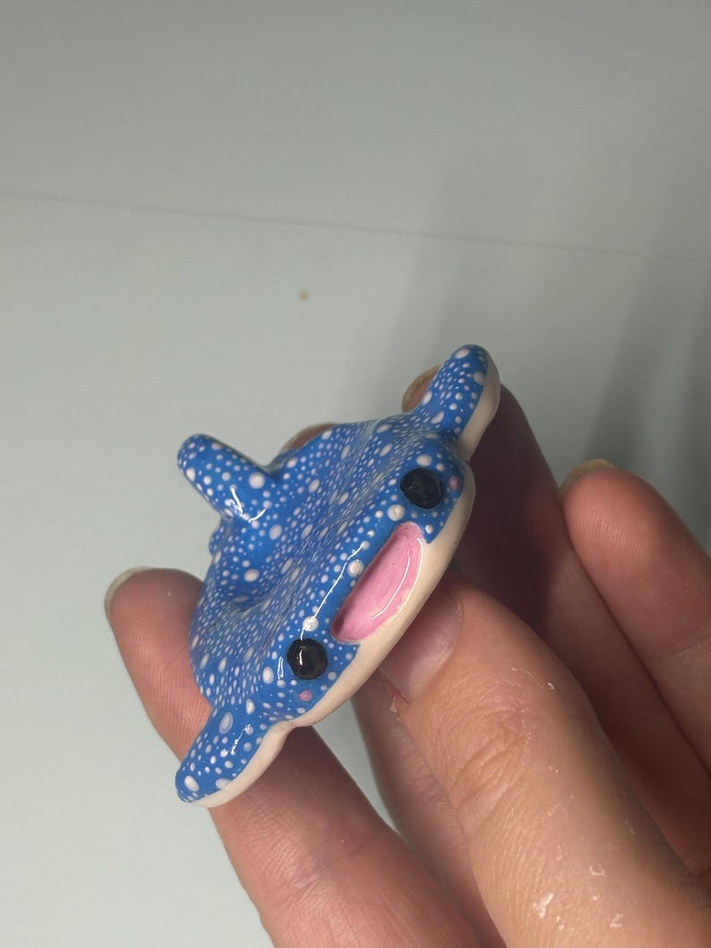 May include: A small, blue whale shark figurine featuring a pattern of white dots across its back, a smooth cream-colored underside, two black circular eyes, and a wide, open mouth with a pink interior. The shark has a dorsal fin on its back.