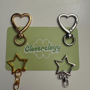 May include: Four metal lobster claw clasps attached to a light green rectangular card with the text Cloverclayy and a clover graphic. Two clasps are heart-shaped in gold and silver tones, and two are star-shaped in gold and silver tones.
