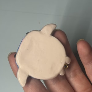May include: A small, smooth, cream-colored ceramic turtle figurine with a rounded shell and four short flippers. The underside is flat and plain, featuring a small gold-toned metal loop attached to one side for hanging or display purposes.