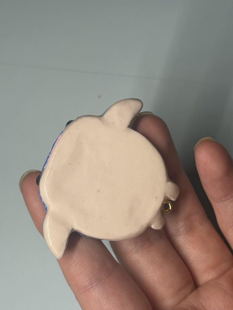 May include: A small, smooth, cream-colored ceramic turtle figurine with a rounded shell and four short flippers. The underside is flat and plain, featuring a small gold-toned metal loop attached to one side for hanging or display purposes.