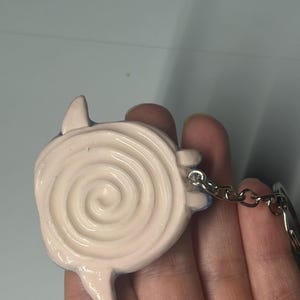 May include: A light pink keychain charm featuring a raised spiral design on the front surface. The charm has small, pointed protrusions extending from the sides and is attached to a silver-toned metal chain and a circular split ring connector.