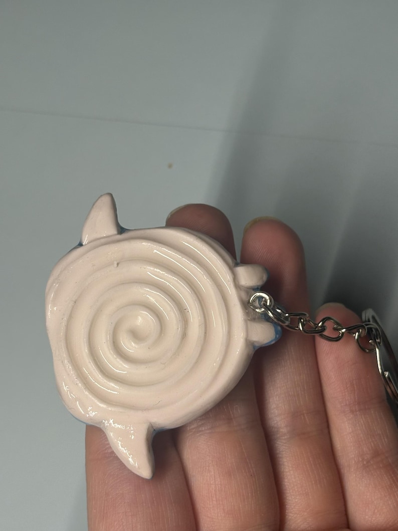 May include: A light pink keychain charm featuring a raised spiral design on the front surface. The charm has small, pointed protrusions extending from the sides and is attached to a silver-toned metal chain and a circular split ring connector.