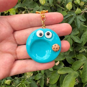 May include: A circular, bright turquoise keyring charm featuring two white eyes with black pupils and a small, tan, brown-spotted cookie attached to the side. The charm is attached to a gold-toned metal lobster clasp and jump ring hardware.