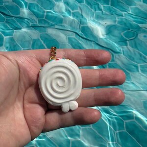 K&ouml;nnte beinhalten: Ein wei&szlig;er, spiralf&ouml;rmiger Lutscher-Anh&auml;nger mit bunten Streuseln am oberen Rand, befestigt an einem goldfarbenen Metallring. Der Anh&auml;nger hat eine glatte, abgerundete Oberfl&auml;che mit einer kleinen, deutlichen Erhebung unten in der Mitte.