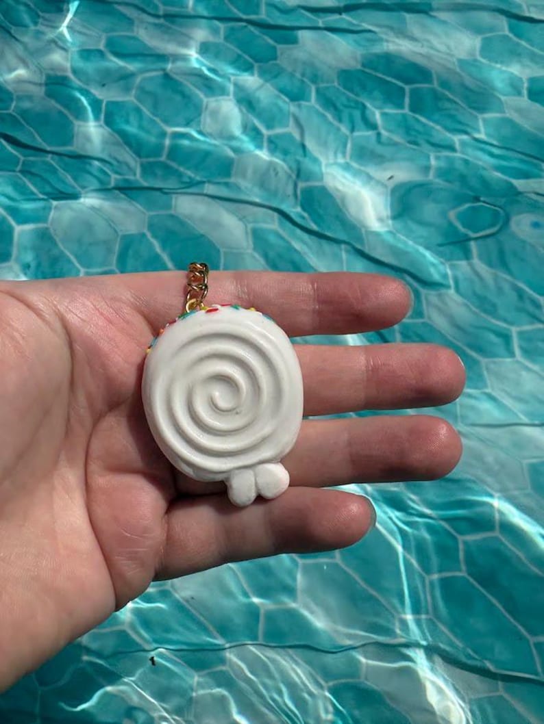 K&ouml;nnte beinhalten: Ein wei&szlig;er, spiralf&ouml;rmiger Lutscher-Anh&auml;nger mit bunten Streuseln am oberen Rand, befestigt an einem goldfarbenen Metallring. Der Anh&auml;nger hat eine glatte, abgerundete Oberfl&auml;che mit einer kleinen, deutlichen Erhebung unten in der Mitte.