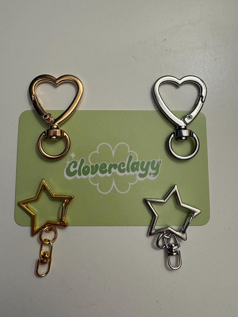 K&ouml;nnte beinhalten: Vier Metall-Karabinerverschl&uuml;sse auf einer hellgr&uuml;nen rechteckigen Karte mit dem Text Cloverclayy. Zwei Verschl&uuml;sse sind herzf&ouml;rmig, einer in Gold und einer in Silber. Zwei sind sternf&ouml;rmig, einer in Gold und einer in Silber, mit kleinem Ring.