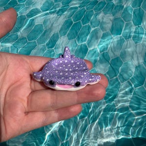 May include: A small, purple whale shark figurine with white polka dots, a smooth texture, and a wide, smiling mouth. The creature features two black, circular eyes and a dorsal fin on its back, designed in a flat, tray-like shape.