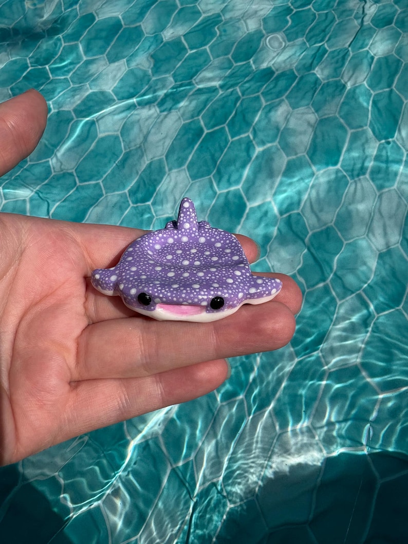 May include: A small, purple whale shark figurine with white polka dots, a smooth texture, and a wide, smiling mouth. The creature features two black, circular eyes and a dorsal fin on its back, designed in a flat, tray-like shape.