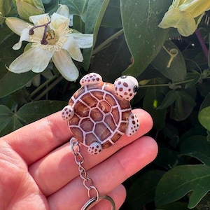 Può includere: Un portachiavi a forma di tartaruga con un guscio marrone dai contorni bianchi e una finitura lucida. La testa e le quattro pinne sono color crema con macchie marroni. Una catenella in metallo argentato e un anello sono fissati alla base.