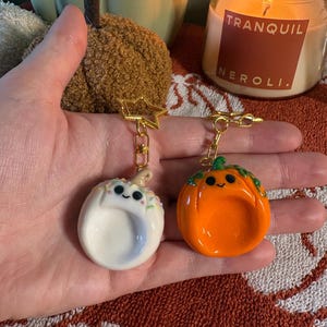 May include: Two pumpkin-shaped keychains with gold-coloured star clasps. One is white with colourful sprinkles and a light brown stem, while the other is bright orange with green vine details. Both feature a smooth, glossy finish and a smiling face.