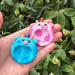 May include: Two circular clay trinket dishes shaped like cow faces. One dish is light blue with darker blue spots, and the other is light pink with darker pink spots. The pink dish features a small strawberry topper, while the blue dish has a blue bead.
