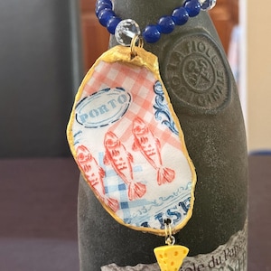 May include: A decorative wine bottle charm featuring a blue and clear beaded loop attached to a gold-rimmed oyster shell. The shell displays a red and white checkered pattern, illustrations of three fish, the word PORTO, and a small yellow cheese charm.