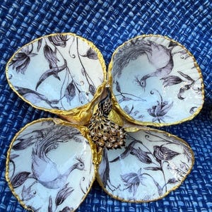 May include: A four-section decorative dish crafted from joined oyster shells, featuring a black and white botanical and peacock print on the interior. The edges are trimmed in gold, and a decorative peacock figurine sits in the center.