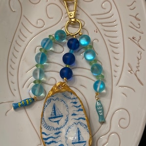 May include: A keyring featuring a gold-tone metal ring and clasp, adorned with blue and green frosted beads and two small blue fish charms. A central oyster shell is painted with blue sailboats and wavy lines, with a gold-painted edge.