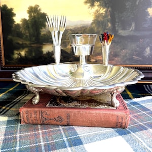 May include: A silver-toned metal serving dish with a scalloped edge and three claw feet, featuring a central pedestal bowl. It is accompanied by a set of silver-toned forks and a small container holding colorful cocktail picks.