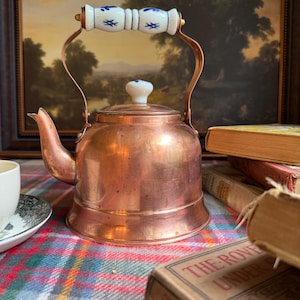May include: A copper tea kettle with a wide base and a curved spout. The handle is made of white ceramic with blue floral patterns, and the lid features a matching white ceramic knob with a small blue floral design at the center.