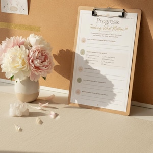May include: A wooden clipboard holding a printed sheet titled Progress: Tracking What Matters with sections for self-reflection. Beside it sits a ceramic vase filled with light pink and white peony flowers and a small cluster of clear crystals.