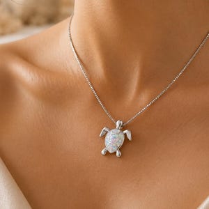 Op de afbeelding: Een zilverkleurige ketting met een zeeschildpadhanger met een iriserende, wit opaalachtige inleg op het schild. De hanger hangt aan een fijne, dunne schakelketting die op het sleutelbeen rust en een gepolijste metalen afwerking heeft.