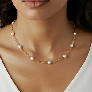 May include: A delicate gold necklace featuring a series of evenly spaced, round, white pearls. The necklace is displayed against a neutral background, highlighting the jewelry's elegant design. The chain is a fine, linked style.