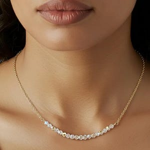 Puede incluir: Un delicado collar dorado con una fila de cristales claros y brillantes. Los cristales están ensartados en una fina cadena dorada, creando un diseño sutil y elegante. El collar se muestra en el cuello de una persona, destacando su delicada naturaleza.