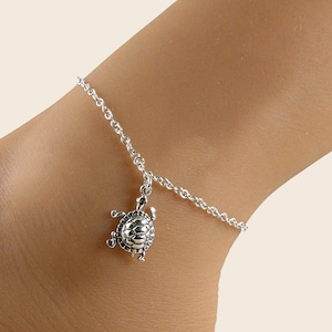 May include: A silver-toned anklet featuring a delicate chain and a small, detailed turtle charm. The charm displays a textured shell pattern and four extended flippers, hanging from a simple link chain that encircles the lower leg area.