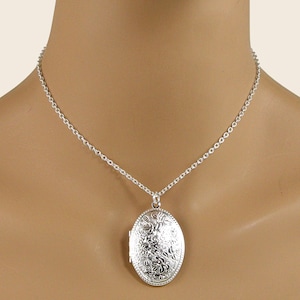 Peut inclure: Un pendentif médaillon ovale de couleur argentée présentant un motif floral en relief complexe sur la surface avant. Le médaillon est suspendu à une délicate chaîne de collier à maillons de couleur argentée autour du cou.