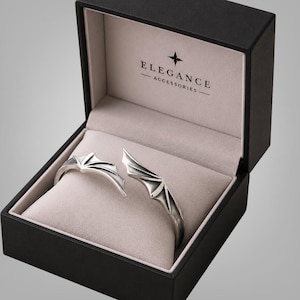 May include: A silver-toned open cuff bracelet featuring two symmetrical bat wing designs, presented inside a black rectangular jewelry box with a light beige interior. The box lid displays the text ELEGANCE ACCESSORIES with a star icon.