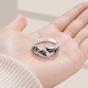 May include: A silver-toned open ring featuring two symmetrical bat wing designs extending from the band. The metal surface has an oxidized finish to highlight the wing details, and the interior of the band is clearly stamped with the text S925.