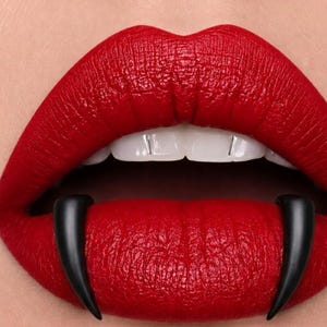 May include: A pair of lips painted with a vibrant matte red lipstick, featuring two long, curved, black fangs extending downward from the upper lip over the lower lip, and two small, thin, vertical silver-colored accents on the upper front teeth.