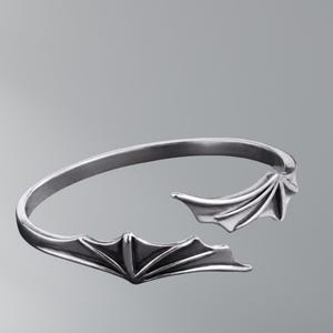 May include: A silver-toned open cuff bracelet featuring two symmetrical bat wing designs at each end. The metal has a polished finish with recessed detailing that accentuates the structure of the wings, creating a continuous curved band shape.