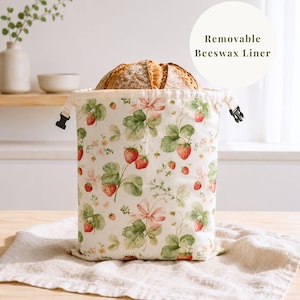 Pode incluir: Um saco de tecido para pão com estampa de morangos e folhas verdes, com fecho de cordão com travas de plástico preto e um forro de cera de abelha removível. O saco contém um pão redondo e tem o texto Removable Beeswax Liner.