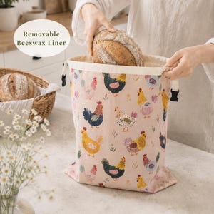 May include: A light pink fabric bread bag featuring a repeating pattern of colorful chickens and small floral sprigs. The bag has a cream-colored top edge with black plastic side buckles and a removable beeswax liner for food storage.