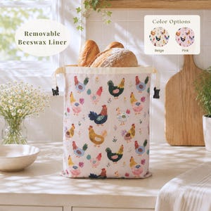 May include: A fabric bread bag with a removable beeswax liner, featuring a repeating pattern of colorful chickens and small flowers. The bag has black side buckles and contains two loaves of bread. Text on the image reads Removable Beeswax Liner.