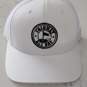 Peut inclure: Une casquette de baseball blanche avec un dos en filet et un panneau avant structuré. Au centre se trouve un écusson brodé noir circulaire représentant un drapeau de golf et le texte FAIRWAY ALL DAY dans une police circulaire.