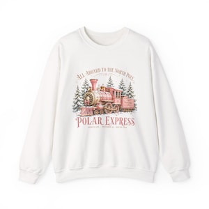 May include: A white crewneck sweatshirt featuring a graphic of a pink steam locomotive surrounded by snow-covered pine trees. The text reads "ALL ABOARD TO THE NORTH POLE", "POLAR EXPRESS", and "ADMITS ONE - DECEMBER 24 - ROUND TRIP".