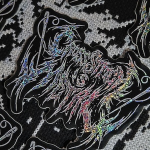 May include: A black sticker featuring the text "CROSS STITCH WITCH" in a stylized, jagged, holographic font. The design includes a needle and thread graphic integrated into the lettering, with a reflective, rainbow-colored finish on the text.
