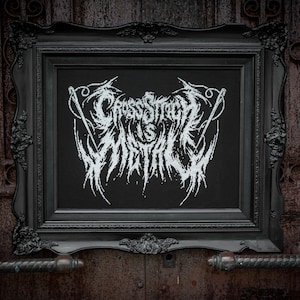 May include: A black rectangular frame containing a cross-stitch embroidery on a black fabric background. The embroidery features white thread forming the text "CROSS STITCH IS METAL" in a jagged, stylized font resembling heavy metal band logos.