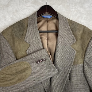 May include: A brown and tan houndstooth blazer featuring olive green suede shoulder yokes and matching elbow patches. The jacket includes a brown satin lining, a notched lapel, and three dark brown buttons on each sleeve cuff. A blue tag reads Made in USA.