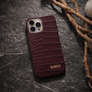 Puede incluir: Una funda para smartphone de color burdeos intenso con un patrón de textura de piel de cocodrilo. La funda presenta un recorte para triple cámara en la parte superior y el texto dorado RM02 grabado en la esquina inferior derecha.