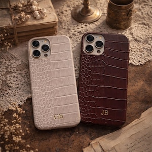 May include: Two smartphone cases with a crocodile skin texture. One case is light beige with the gold embossed initials GB, and the other is dark brown with the gold embossed initials JB. Both cases feature a triple camera cutout on the back.