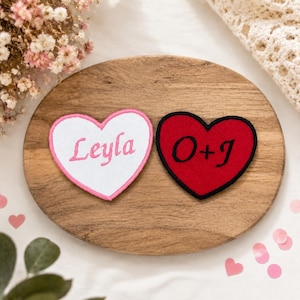 May include: Two heart-shaped embroidered patches. The left patch is white with a pink border and the name Leyla embroidered in pink script. The right patch is red with a black border and the text O+J embroidered in black script.