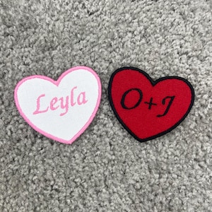 May include: Two heart-shaped embroidered patches. The left patch is white with a pink border and the name Leyla embroidered in pink. The right patch is red with a black border and the text O+J embroidered in black script lettering.