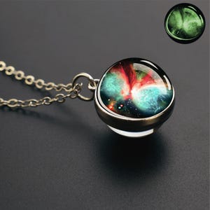 May include: A silver-toned necklace featuring a round glass pendant with a vibrant nebula design in shades of teal, red, and black. The pendant displays a glowing effect in the upper right corner, showcasing a bright green luminescence against a dark background.