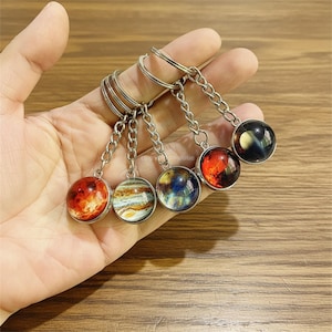 Könnte beinhalten: Ein Set aus vier silberfarbenen Metallschlüsselanhängern, die jeweils mit einem runden Glaskabochon-Anhänger versehen sind. Die Anhänger zeigen detaillierte, farbenfrohe Himmelsbilder, die Planeten und Nebeln in Rot-, Orange-, Blau- und Schwarztönen ähneln.