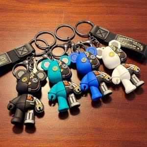 May include: Four bear-shaped keychains featuring a split design with one side in solid black, teal, blue, or white, and the other side in a black mechanical pattern with gear details. Each keychain includes a metal ring and a black fabric strap.