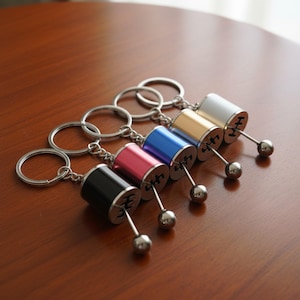 May include: Five metal gear shift keychains arranged in a row, featuring cylindrical bodies in black, pink, blue, gold, and silver. Each keychain includes a circular split ring, a short metal stick with a spherical tip, and a gear pattern engraving.