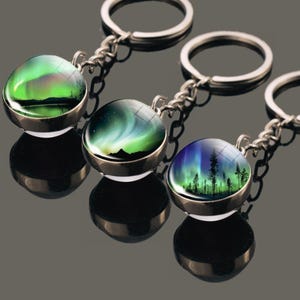 Op de afbeelding: Drie ronde sleutelhangers met koepelvormige glazen cabochons met levendige afbeeldingen van het noorderlicht. Elke sleutelhanger heeft een zilverkleurige metalen zetting, een korte ketting en een ring voor sleutels, met groene en paarse lichteffecten.