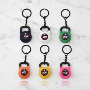 Puede incluir: Seis llaveros con forma de pesa rusa con anillas y cadenas de metal negro. Las pesas son azul marino, blancas, verdes, naranjas, amarillas y rosas. Cada una tiene un emblema circular central con una estrella blanca y un número: 700, 200, 200, 500, 500 o 400.