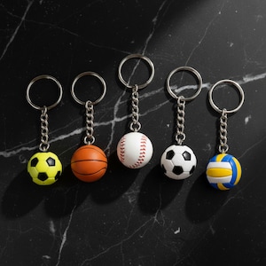 以下が含まれることがあります： ネオンイエローと黒のサッカーボール、オレンジのバスケットボール、赤いステッチの白い野球ボール、白黒のサッカーボール、青・黄・白のバレーボールなど、ミニチュアのスポーツボールが付いた5つの金属製キーホルダーのセット。